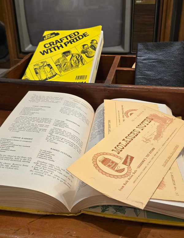 An open vintage book on a wooden table with a yellow 'Crafted with Pride' book resting on top and two tan certificates or cards placed over the pages. A vintage television set is visible in the background.