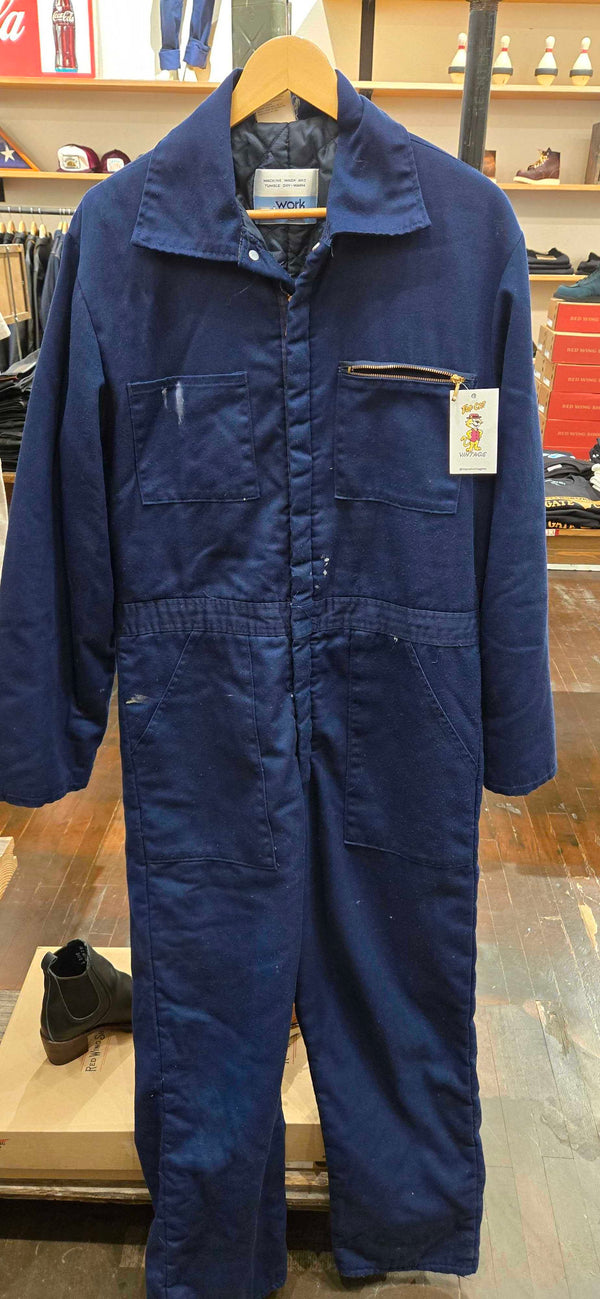 A dark navy blue work jumpsuit hanging on a wooden hanger in a retail store. The jumpsuit has a high collar, front button closure, and multiple utility pockets.