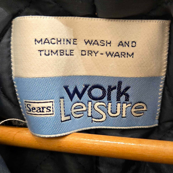 Close-up of a sewn-on label reading 'MACHINE WASH AND TUMBLE DRY-WARM' with 'work Leisure' and a Sears logo, attached to a dark quilted jacket on a wooden hanger.