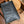 A black textured leather wallet or clutch with a gold-toned edge, resting on a rustic wooden shelf next to a round wooden coaster.