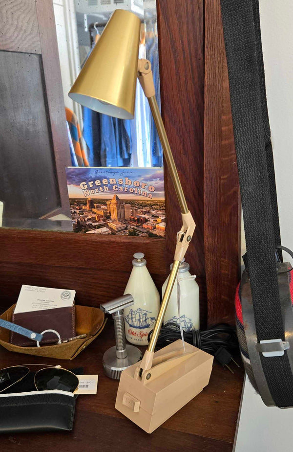 A vintage adjustable desk lamp with a gold metal arm and a light wood base, sitting on a dark wooden desk next to other household items.