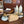 A wooden desk or dresser surface with a brass and wood adjustable desk lamp in the foreground, alongside a bowl, bottles, and other household items in a real indoor setting.