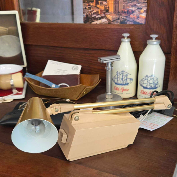A wooden desk or dresser surface with a brass and wood adjustable desk lamp in the foreground, alongside a bowl, bottles, and other household items in a real indoor setting.