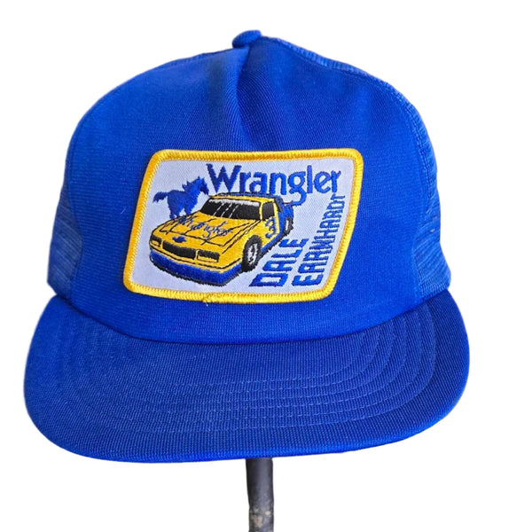 A bright blue Wrangler trucker hat with a yellow and blue patch on the front, displayed on a stand inside a retail store.